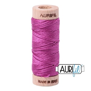 May include: A spool of vibrant magenta sewing thread wrapped around a cylindrical wooden core. The top of the wooden spool is branded with the text AURIFIL, while the bottom features the text MADE IN ITALY. The thread has a visible sheen.