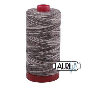 May include: A cylindrical spool of thread featuring a variegated pattern of dark gray, light gray, and white fibers. The spool has a solid red plastic base and a matching red top cap. The brand name AURIfil is printed in black and blue text.
