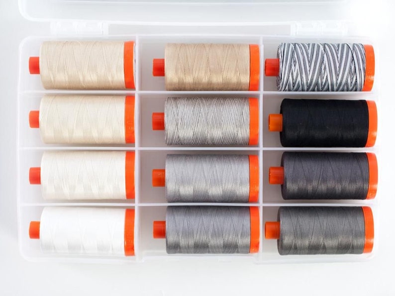 Op de afbeelding: Een transparante kunststof opbergdoos met 12 spoelen garen. De spoelen zijn gerangschikt in drie rijen van vier. De spoelen zijn in tinten beige, grijs en zwart.
