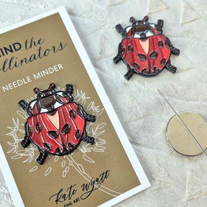 May include: A red and black ladybug needle minder with a matching pin. The needle minder is on a card that reads "Mind the Pollinators" and "Needle Minder" with the artist's signature. A silver needle and a round magnet are also in the image.