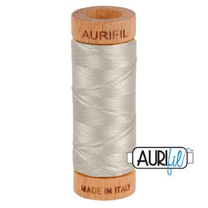 May include: A spool of light gray sewing thread wound around a cylindrical wooden core. The top wooden section is branded with the text AURIFIL, and the bottom wooden section features the text MADE IN ITALY. The Aurifil logo is visible.