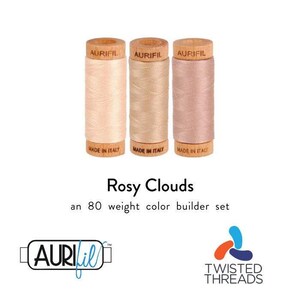 May include: Three spools of Aurifil thread in shades of light pink, beige, and dusty rose. Each wooden spool is labeled Made in Italy. The text Rosy Clouds an 80 weight color builder set is displayed below the spools, along with the Aurifil logo.