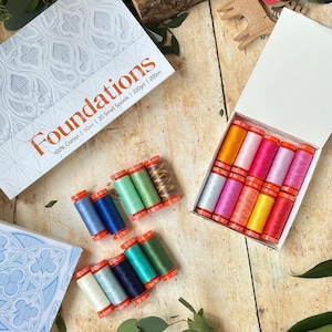 May include: A set of Foundations 100% cotton thread spools, 50wt, 20 small spools, 220yd, 200m. The collection includes a white box with a blue geometric pattern and an open box containing ten colorful thread spools, with ten additional spools arranged outside.