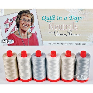 May include: Six large spools of 50wt 100% cotton thread in neutral shades, each with a red cap and base. The packaging features the text Quilt in a Day Neutrals by Eleanor Burns and specifies 1422 yards per spool, with the Aurifil brand logo.