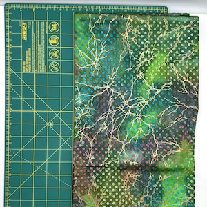May include: A folded fabric with a green, blue, and purple spotted pattern. The fabric has a gold, branch-like design overlaying the spotted pattern. The fabric is next to a green cutting mat with measurement markings.