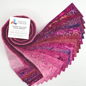 May include: A roll of fabric strips in shades of pink and purple, fanned out to display the various patterns and colors. The fabric has a batik-style design. A white card with the text "THANK YOU FOR YOUR ORDER!" is visible.