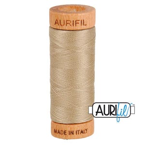 May include: A spool of light brown sewing thread wound around a wooden core. The top of the wooden spool is branded with the text AURIFIL, and the bottom is branded with the text MADE IN ITALY. A logo with the text AURIFIL is visible on the right.