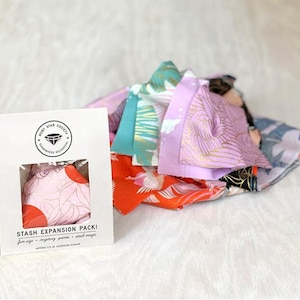 May include: A white card with the text "STASH EXPANSION PACK!" and a window showing a pink fabric bundle with a floral design. Assorted fabric scraps in various colors and patterns are scattered around the card, including orange, teal, and purple fabrics.