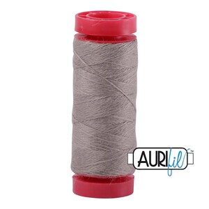 May include: A spool of light brown sewing thread wound onto a cylindrical plastic core with bright red caps at the top and bottom. The brand name AURIfil is printed in black and blue text on a white rectangular label in the lower right corner.