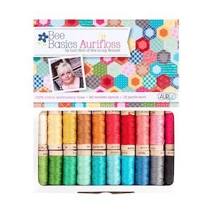 May include: A set of 20 wooden spools of 100% cotton embroidery floss, each containing 18 yards, presented in a box labeled Bee Basics Aurifloss by Lori Holt. The packaging features a colorful hexagonal pattern and a photograph of the designer.