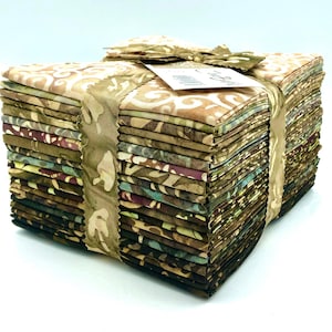 May include: A stack of folded fabric squares secured with a tan ribbon featuring a leaf pattern. The fabric pieces display various batik-style prints in earthy tones including beige, brown, olive green, and deep purple, with a white product tag.