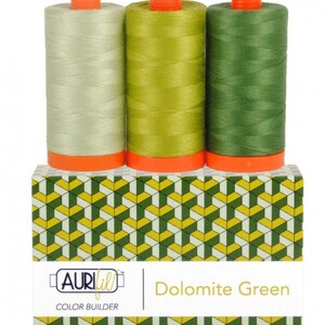 May include: Three spools of sewing thread in light, medium, and dark green shades, each with orange caps and bases. They sit atop a rectangular box featuring a geometric pattern of green, yellow, and white, labeled Aurifil Color Builder Dolomite Green.
