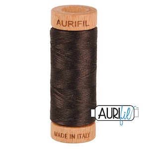 May include: A spool of dark brown thread featuring a light-colored wooden top and bottom. The top wooden piece is embossed with the text AURIFIL, and the bottom wooden piece is embossed with the text MADE IN ITALY. The thread is wound tightly.