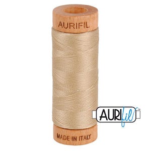 May include: A spool of light beige sewing thread wound around a wooden core. The top of the wooden spool is branded with the text AURIFIL, and the bottom is branded with the text MADE IN ITALY. The thread has a smooth, consistent texture.