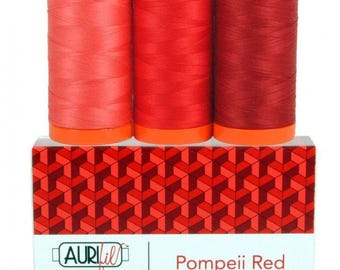 May include: Three spools of thread in varying shades of red, ranging from light coral to deep crimson, sit atop a rectangular box. The box features a geometric pattern in red tones and a white label with the text Aurifil Color Builder Pompeii Red.