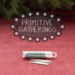 May include: A clear plastic tube with a white label that reads "Bag Stitch Quilting Needles". The tube contains a set of silver quilting needles. The tube is sitting on a red surface. The image is labeled "Primitive Gatherings".