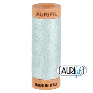 May include: A spool of light blue sewing thread wound around a wooden cylinder. The top of the wooden spool is branded with the text AURIFIL, and the bottom is branded with the text MADE IN ITALY. The thread has a smooth, consistent texture.