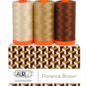 May include: Three spools of sewing thread in light beige, medium tan, and dark brown, each with an orange cap and base. They sit atop a rectangular box featuring a geometric brown and white pattern, labeled Aurifil Color Builder Florence Brown.