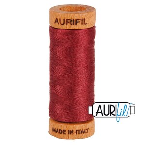 May include: A spool of deep red sewing thread wound around a cylindrical wooden core. The top of the wooden spool is branded with the text AURIFIL, and the bottom is branded with the text MADE IN ITALY. The thread has a smooth, matte finish.
