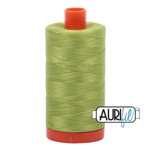 May include: A spool of light olive green sewing thread with a bright orange plastic top and base. The spool features the brand name Aurifil printed in black and blue text on a white rectangular label attached to the lower section of the thread.