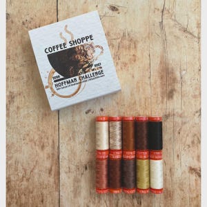 May include: A white square box labeled COFFEE SHOPPE HOFFMAN CHALLENGE 2026 2027 with a coffee cup graphic, accompanied by ten spools of thread in various shades of brown, cream, and black, each featuring orange labels with 50wt and 200m text.