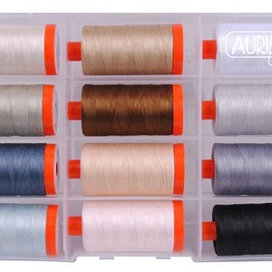 May include: A clear plastic storage case containing twelve spools of thread, each with bright orange end caps. The spools feature various neutral and dark colors including white, beige, brown, gray, blue, and black. The Aurifil logo is printed in white.