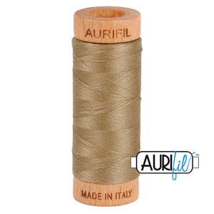May include: A spool of Aurifil thread in a light brown color. The spool is made of wood and has the Aurifil logo printed on it. The thread is made in Italy.