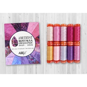 May include: A collection of Aurifil cotton thread spools in various shades of pink, purple, and gold, alongside a promotional card for the Amethyst Hoffman Challenge 2025-2026. The card features a geometric design and product details.