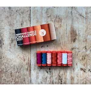 May include: A set of seven small spools of thread in a rectangular box labeled Windham Fabrics Challenge 2027. The spools feature a gradient of colors including black, red, orange, and beige. Below the box are seven individual spools in purple, blue, pink, orange, red, white, and variegated pink.