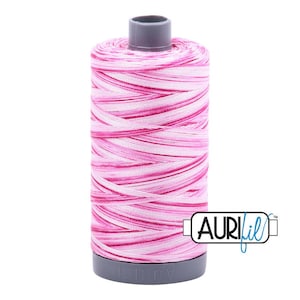 May include: A spool of sewing thread featuring a variegated color pattern of alternating light pink, medium pink, and white strands wound around a grey plastic core. The brand name AURIfil is printed in black text on the bottom grey base.