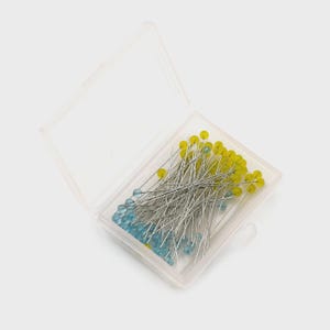 May include: A clear plastic box containing sewing pins. The pins have silver shafts and colorful heads, including yellow and blue. The box is open, revealing the contents inside. The pins are likely used for crafting or sewing projects.