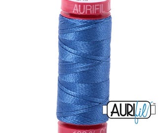 May include: A spool of blue sewing thread with a red plastic top and bottom. The top label reads AURIFIL, Cotton 12, and 6738. The bottom label reads 100% Cotton. A logo with the text AURIFIL is positioned to the right of the spool.