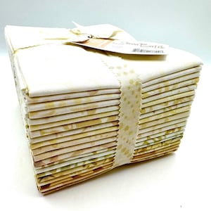 May include: A stack of folded fabric squares featuring a cream base with subtle yellow and green batik patterns. The stack is secured with a cream-colored ribbon printed with yellow dots and includes a tag labeled Java Batik.