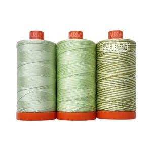 May include: Three spools of Aurifil sewing thread with orange plastic bases and caps. The left spool is light sage green, the center is a medium mint green, and the right spool features a variegated pattern of white, light green, and dark olive green.