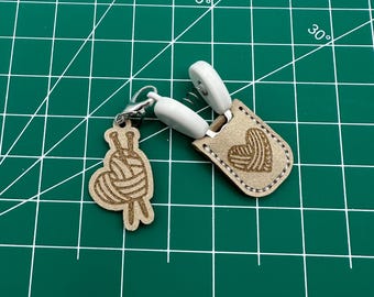 May include: A set of two white and brown yarn needle holders. The holders are shaped like hearts and feature a yarn ball and knitting needles design.