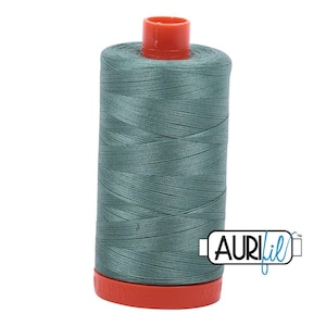 May include: A spool of sage green sewing thread featuring a bright orange plastic top and base. The spool is wrapped in smooth, tightly wound thread and displays the AuriFil brand logo in black and blue text on the lower right side of the image.