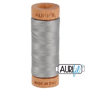 May include: A spool of gray Aurifil thread. The thread is wound around a cylindrical core with wooden ends. The top wooden end has the brand name "AURIFIL" engraved. The bottom wooden end reads "MADE IN ITALY". The Aurifil logo is on the right.