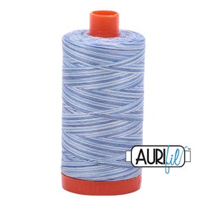 May include: A spool of sewing thread featuring a variegated pattern of light blue, medium blue, and white horizontal stripes. The spool has a bright orange plastic top and base, with the brand name AURIfil printed in black and blue text.