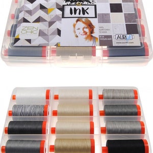 Op de afbeelding: Een transparante kunststof opbergdoos gevuld met 12 spoelen Aurifil garen. De spoelen zijn in verschillende tinten grijs, zwart en beige. De doos heeft een label met de tekst "Paper Ink" en "Zen Chic".