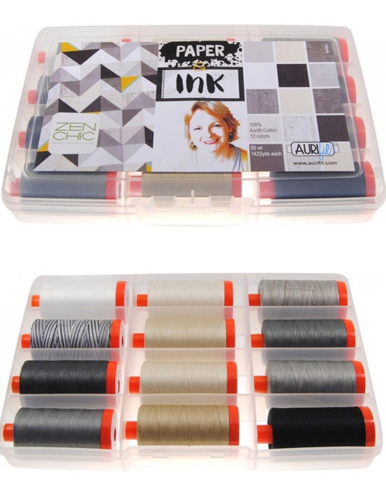 Op de afbeelding: Een transparante kunststof opbergdoos gevuld met 12 spoelen Aurifil garen. De spoelen zijn in verschillende tinten grijs, zwart en beige. De doos heeft een label met de tekst "Paper Ink" en "Zen Chic".