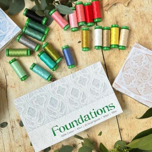 May include: A collection of twenty colorful sewing thread spools in shades of green, pink, red, yellow, blue, and grey, arranged near a white box labeled Foundations 100% Cotton 40wt 20 Small Spools 164yd 150m with a decorative pattern.