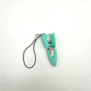 May include: A small, teal-colored phone clip with a black lanyard. The clip has a spring mechanism and a silver-colored metal accent. The lanyard is a thin, black cord.