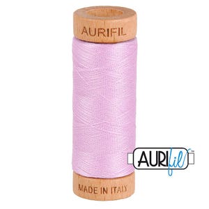 May include: A spool of light pink sewing thread wrapped around a cylindrical wooden core. The top of the wooden spool is embossed with the text AURIFIL, and the bottom is embossed with the text MADE IN ITALY. The Aurifil logo is visible.