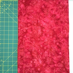 May include: A piece of red fabric with a floral pattern. The fabric is a deep red color with lighter red floral designs. The fabric is next to a green cutting mat with yellow grid lines and measurements in inches.