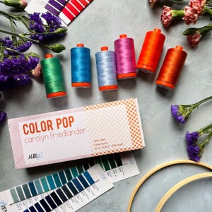 May include: Six spools of 50wt cotton thread in green, blue, light blue, pink, orange, and red, accompanied by a rectangular box labeled Color Pop by Carolyn Friedlander, Aurifil, 100% cotton, 6 large spools, 1422yd/1300m each, and color charts.