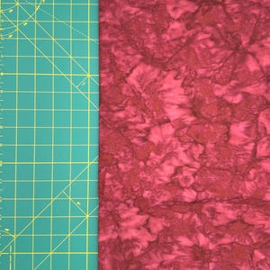 May include: A section of red fabric with a mottled pattern, next to a green cutting mat with yellow grid lines and inch measurements. The fabric has a textured appearance with darker red and brown accents.