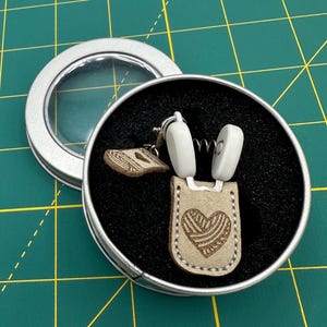May include: A pair of white yarn bowl holders with a spring mechanism. The holders are in a round tin with a black interior and a clear lid. The holders have a beige leather strap with a heart-shaped yarn ball design.