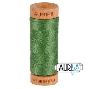 May include: A spool of green sewing thread wrapped around a wooden core. The top of the wooden spool is branded with the text AURIFIL, and the bottom is branded with the text MADE IN ITALY. The thread is a solid, medium-toned green color.