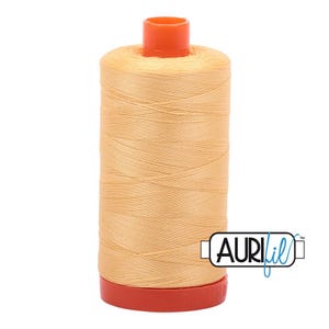 May include: A spool of light orange sewing thread wound around a cylindrical core with a bright orange top and bottom cap. The brand name Aurifil is printed in black and blue text on a white rectangular label attached to the side of the spool.