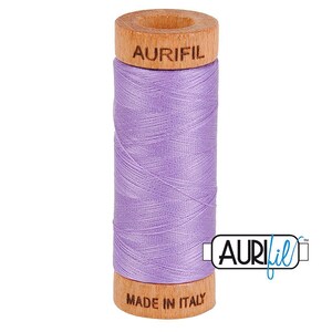 May include: A spool of light purple sewing thread wound around a cylindrical wooden core. The top of the wooden spool is branded with the text AURIFIL, and the bottom is branded with the text MADE IN ITALY. The thread has a smooth texture.
